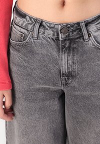 Pelēki džinsi no denima ar izbalinātu faktūru, ar metāla pogu aizdari un divām priekšējām kabatām, novietoti virs sarkana garām piedurknēm krekla.