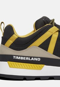 Zwarte en gele sneaker met een gestructureerde bovenkant, voorzien van een versterkte hiel en contrasterende accenten. Merkennaam "TIMBERLAND" in het wit.