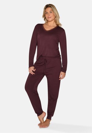 Femme debout pieds nus portant un haut à manches longues et un pantalon assortis en tenue d'intérieur bordeaux foncé, posant avec une jambe légèrement pliée.