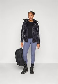 Svart vadderad jacka med dragkedja, svart långärmad topp, grå leggings, svarta vandringsskor och en mörk ryggsäck. Släta och eleganta texturer.