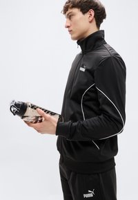 Μαύρο αθλητικό μπουφάν με ψηλό γιακά, λευκές ραφές και φερμουάρ, κρατημένο από ένα άτομο που κρατάει ένα διαφανές, διάφανο μπουκάλι νερού.