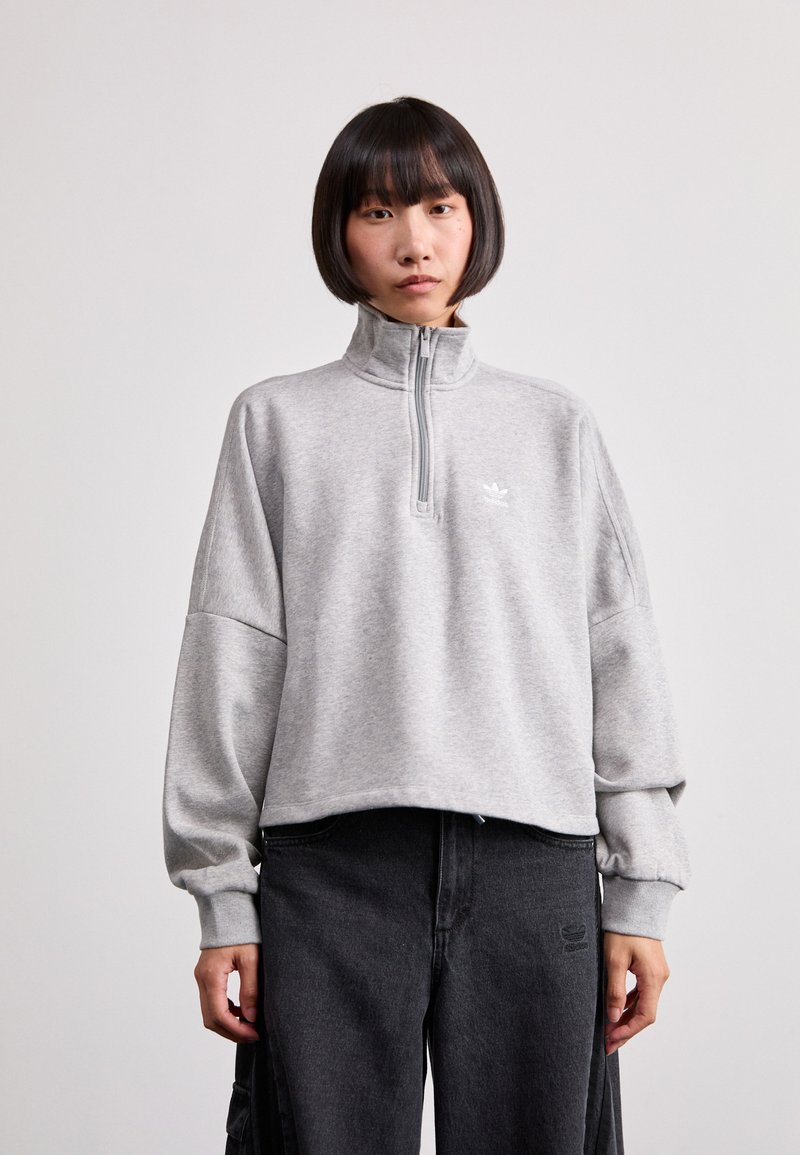Sudadera gris con cremallera, hecha de un material suave, con un alto cuello, hombros caídos y un pequeño logo blanco en el pecho.