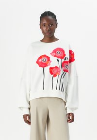 Felpa bianca con fiori rossi ricamati e steli neri, spalle cadenti e vestibilità rilassata. Abbinata a pantaloni beige.