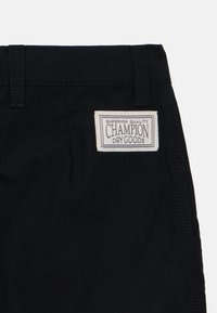 Svart shorts med vevet tekstur, med en hvit merkelapp med teksten "Champion Dry Goods" og kontrasterende sømmer på livet.