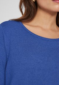 Blå stickad tröja med vid halsringning. Texturen är mjuk och ribbad, med en solid färg utan mönster eller utsmyckningar.