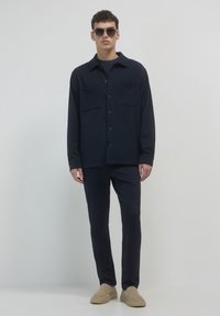 Giacca camicia blu navy con due tasche sul petto, abbinata a pantaloni coordinati e scarpe beige slip-on, su uno sfondo neutro.