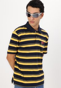 Gul og marineblå stribet polo skjorte med krave, der har et rødt logo patch. Stoffet ser glat ud, med korte ærmer og en afslappet pasform.