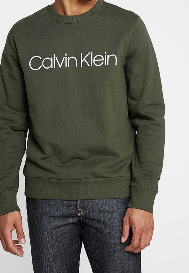 Felpa verde in cotone con scollatura a girocollo, polsini a costine e logo bianco "Calvin Klein" sul petto. Abbinata a jeans scuri.