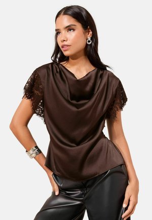 Žena oblečená v tmavohnedom saténovom topánke s krátkymi čipkovanými rukávmi a čiernych kožených nohaviciach, doplnená striebornými náušnicami a náramkom.