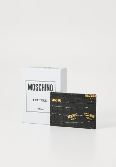 Črni usnjeni držalec za kartice s krokodilskim vzorcem, opremljen z zlato "Moschino" blagovno znamko, predstavljen ob belem škatli z minimalističnim dizajnom.