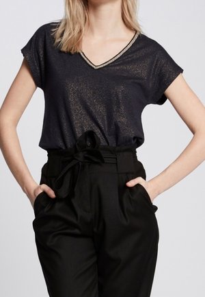 Woman wearing a shimmery black V-neck short sleeve top tucked into high-waisted black pants with a matching fabric belt tied in a bow.