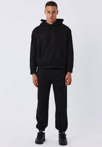 Sudadera negra con bolsillo canguro y capucha ajustable, combinada con pantalones de chándal ajustados a juego. Ambos artículos cuentan con una tela suave y texturizada.