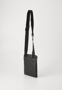 Sac en cuir noir à bandoulière avec une finition texturée, doté d'une fermeture éclair prononcée, d'une sangle en nylon réglable et d'une forme rectangulaire mince.