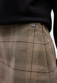 Gonna a quadri marrone con una combinazione di linee sottili e spesse. Include una chiusura con zip e un piccolo logo ricamato in verde e rosa.
