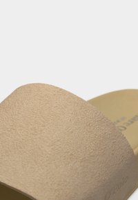 Σανδάλι από μπεζ σουέντ με φαρδιά λουρί πάνω από το πόδι και επίπεδη σόλα, εμφανίζεται από κοντινή γωνία.