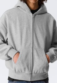 Sweat à capuche gris zippé avec une poche kangourou, des poignets et un ourlet côtelés. Texture douce et polaire ; coupe décontractée. Le design comprend une capuche et des détails zippés.