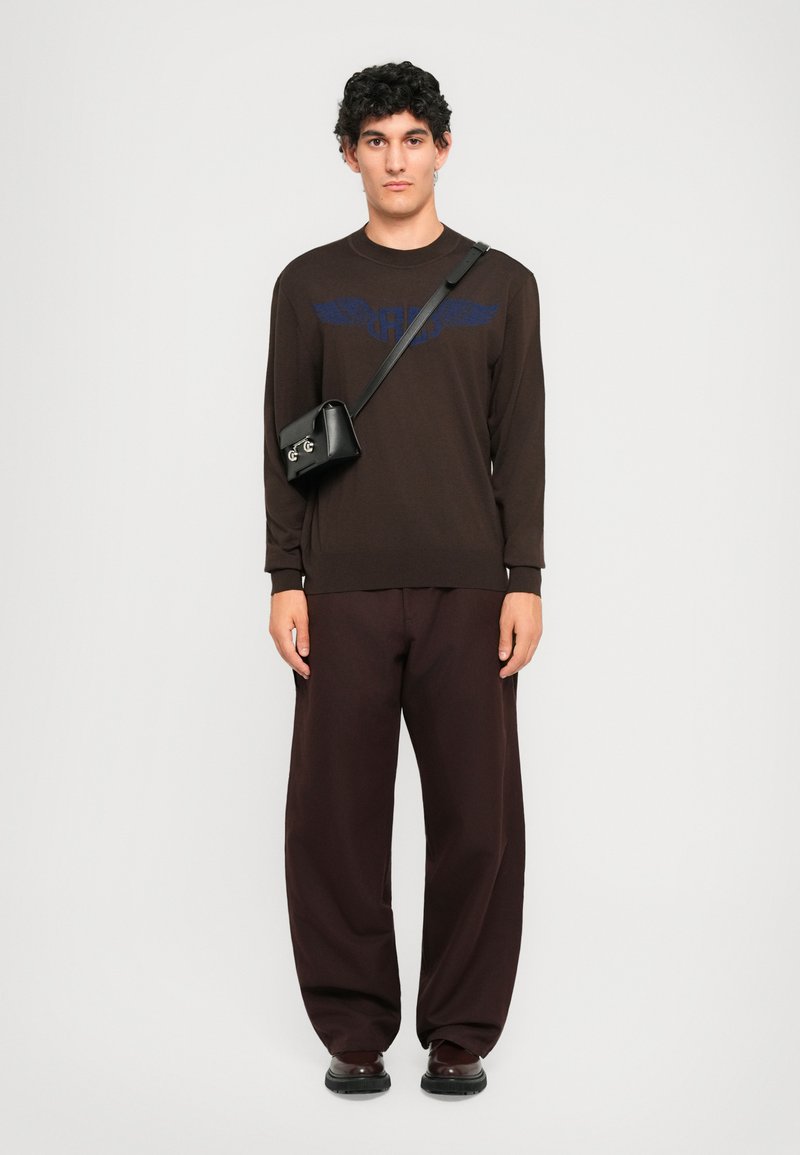 Un jeune homme aux cheveux bouclés portant un pull marron foncé, un pantalon large, un sac bandoulière noir et des chaussures foncées, se tenant devant un fond blanc.