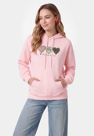 Ung kvinde med langt bølget hår bærer en lyserød hættetrøje med tre hjerte-designs og tekst, kombineret med lyseblå jeans og røde hjerte-øreringe.