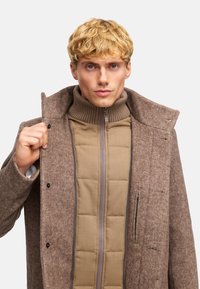 Abrigo de mezcla de lana marrón con cuello alto, que presenta una chaqueta interior beige con cremallera. Tejido texturizado y detalle de botones a presión.