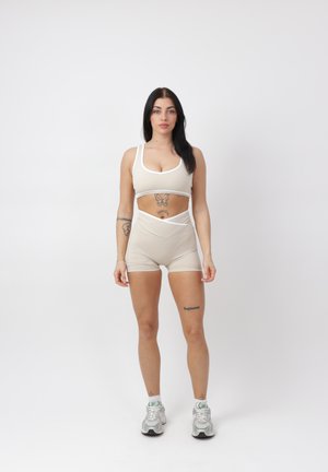 Frau mit langen dunklen Haaren, die einen beigen Sport-BH und Shorts trägt, steht vor einem schlichten weißen Hintergrund mit neutralem Ausdruck.