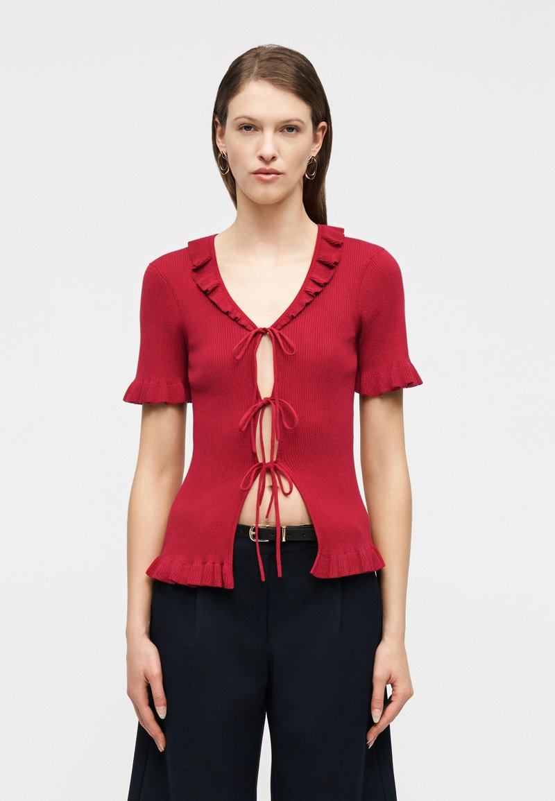 Mujer con un top de punto rojo de manga corta con bordes ondulados y tres lazos delanteros, combinado con pantalones negros y pendientes de aro dorados.