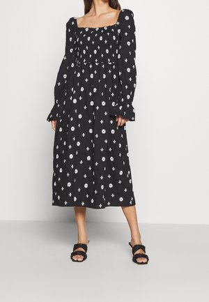 Vrouw draagt een zwarte midi-jurk met witte bloem- en kruismotieven, lange pofmouwen, een vierkante halslijn en zwarte sandalen met gevlochten bandjes.