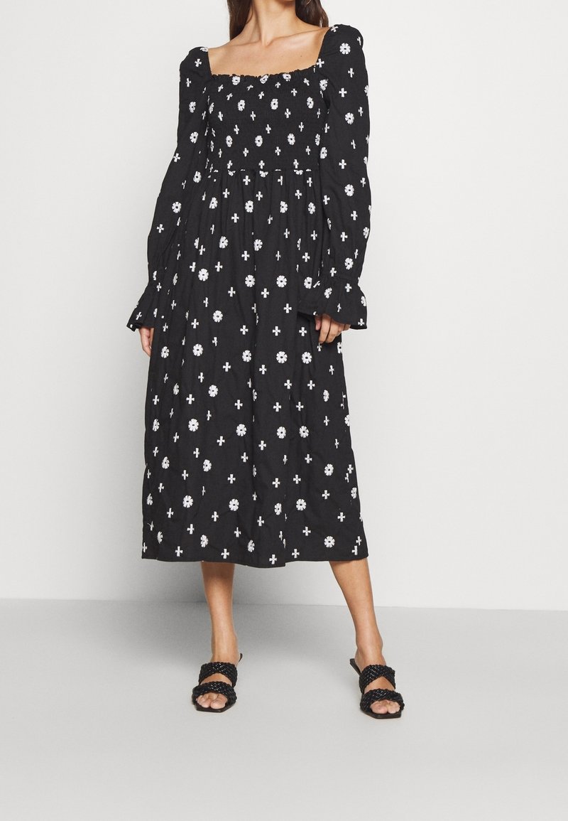 Femme portant une robe midi noire avec des motifs floraux blancs et des croix, des manches longues bouffantes, un décolleté carré et des sandales noires à brides tressées.