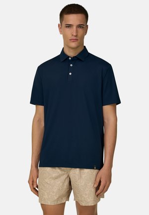 Junger Mann trägt ein marineblaues kurzärmeliges Polohemd und beige gemusterte Shorts, steht vor einem einfarbigen hellen Hintergrund.