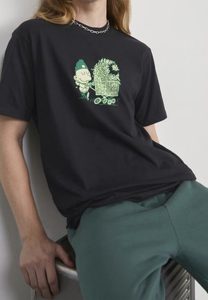 Sort bomulds t-shirt med et grønt tegneserie motiv af en person, der skubber en vogn fyldt med blomster. Model sidder ned iført grønne bukser.