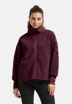 Jeune femme aux cheveux attachés, portant une veste polaire texturée violet foncé à col haut zippée et un pantalon noir sur un fond uni.