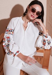 Witte button-up overhemd met kleurrijke bloemenborduursels op pofy lange mouwen; gedragen met witte broek; model zit op een houten stoel.