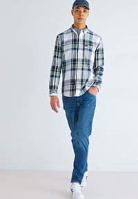 Camisa de cuadros en verde, azul marino y blanco con mangas largas, combinada con jeans azules y zapatillas blancas. El modelo lleva una gorra.