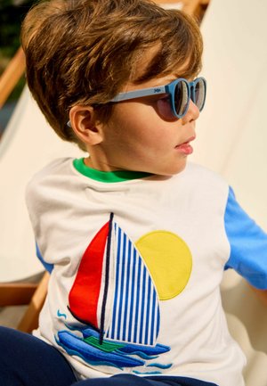 Mladý chlapec s modrými slunečními brýlemi a bílým tričkem s barevným motivem plachetnice a slunce, sedící na dřevěné židli venku.