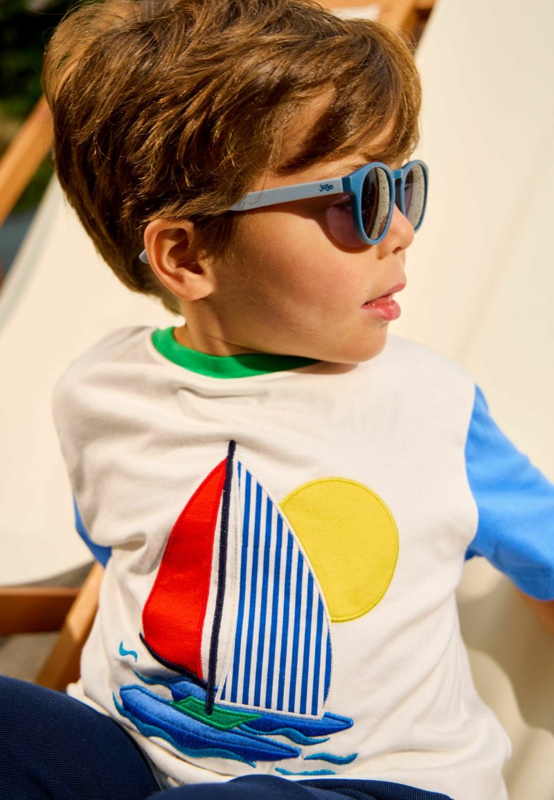Young boy wearing blue sunglasses and a white shirt with a colorful sailboat and sun design, seated in a wooden chair outdoors.