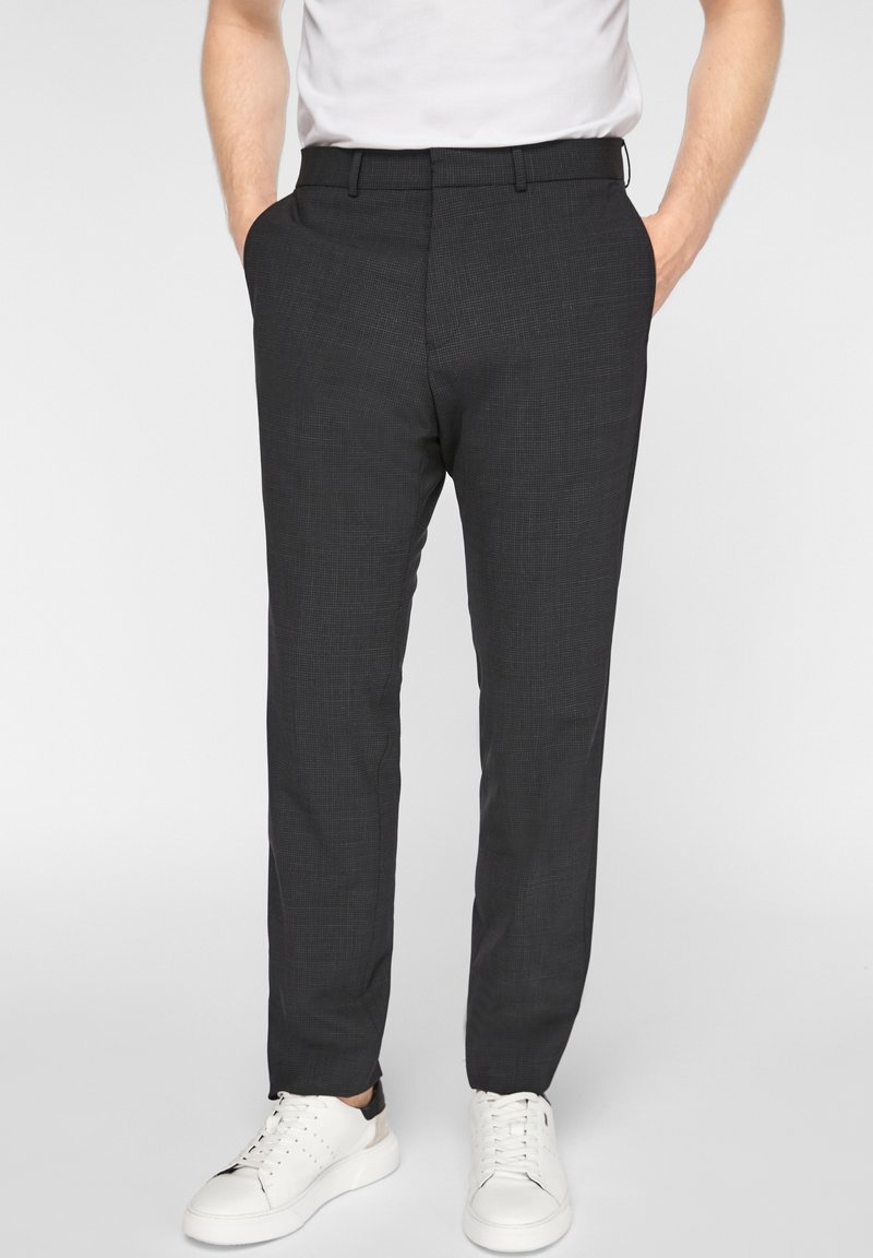 Homme portant un pantalon slim gris foncé avec des poches latérales et des baskets blanches, debout devant un fond clair uni.