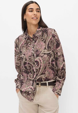 Kvinde iført en brun skjorte med paisley-mønster og lange ærmer, stukket ned i beige bukser med sort bælte, hænderne i lommerne, ser til siden.