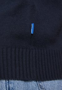 Pletený námořnický svetr s ribbovaným lemem a připojenou modrou nálepkou, vystavený na světlé denimové tkanině. Celkově hladká textura.