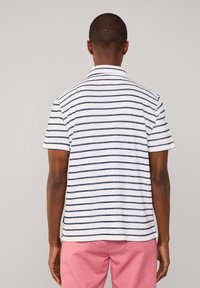 Una polo bianca con righe orizzontali blu navy, maniche corte e colletto classico, abbinata a pantaloni rosa, vista da dietro.