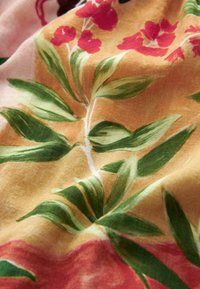 Tissu doux avec des feuilles vertes et des fleurs rouges imprimées sur un fond jaune chaud et rose dans une disposition fluide.
