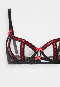 Čierna sieťovaná podprsenka s červenými akcentmi, s ozdobnými vlnkovanými okrajmi, dekoratívnymi mašľami a štruktúrovanou kostrou pre podporu a tvar.