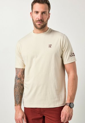 Mann mit Bart und tätowiertem linkem Arm, trägt ein beiges JP 1880 T-Shirt mit Logo auf Brust und Ärmel, rote Hose und eine schwarze Armbanduhr am rechten Arm.