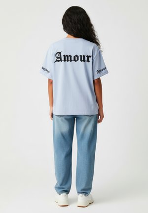 Lichtblauwe oversized t-shirt met zwarte "Amour" tekst op de achterkant en mouwen, gecombineerd met loszittende vervaagde blauwe jeans en witte sneakers.