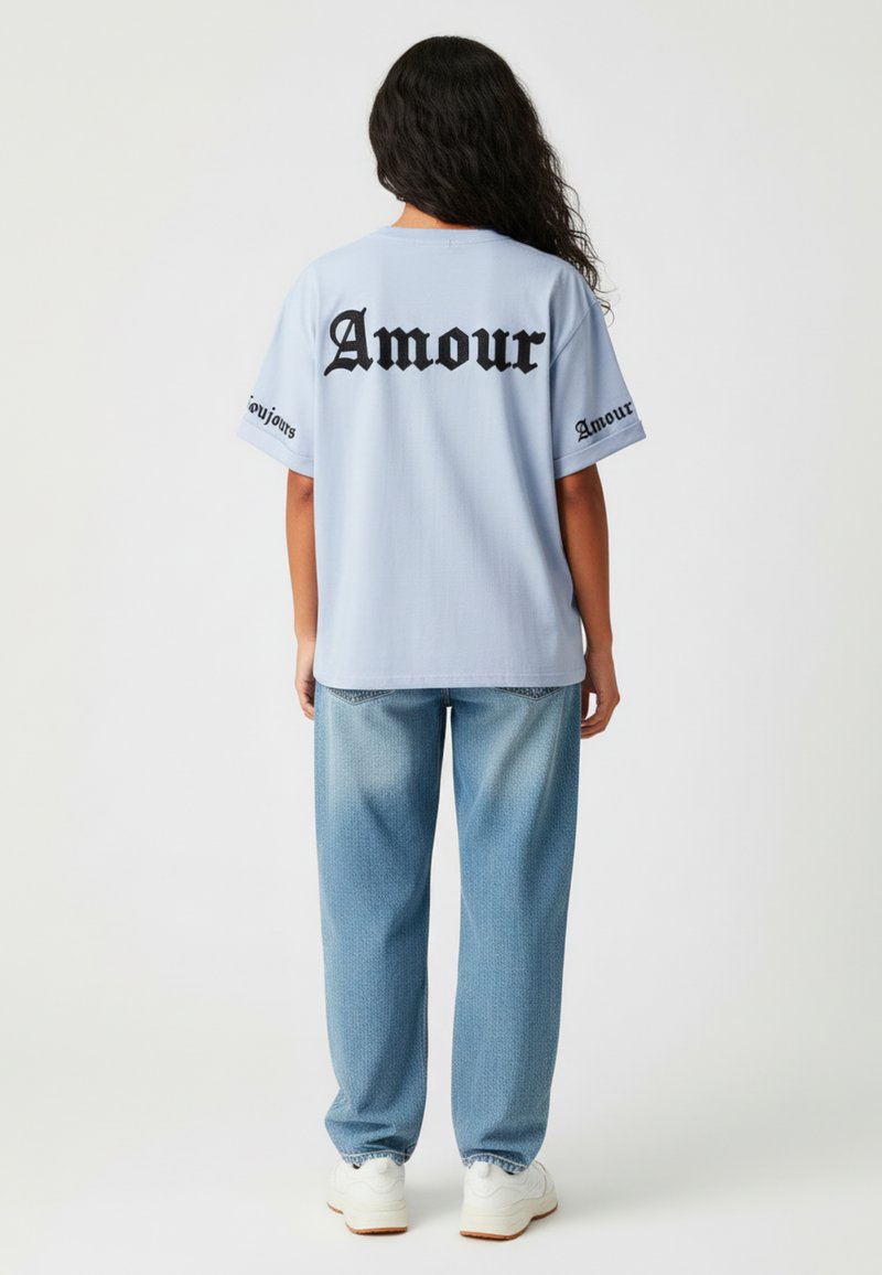 T-shirt oversize bleu clair avec le texte noir "Amour" dans le dos et sur les manches, associé à un jean bleu clair ample et des baskets blanches.