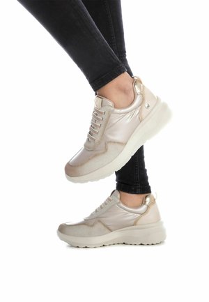 Zapatillas color beige claro con detalles metálicos suaves, paneles texturizados y una suela blanca gruesa. El sistema de cordones cuenta con cinco ojales.