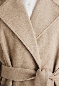 Abrigo de lana beige con cuello de muesca, solapas y cinturón de lazo; presenta una textura suave y un detalle de botón para el cierre.