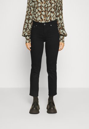 Jeans de mezclilla negra con un diseño ajustado, cinco bolsillos y un cierre clásico de botones, combinados con una blusa de manga larga con un patrón y zapatos oscuros.