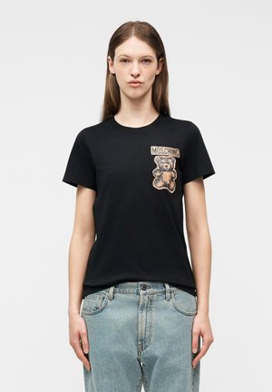 Γυναίκα που φοράει μαύρο μπλουζάκι Moschino με ένα σχέδιο αρκουδάκι στο στήθος, συνδυασμένο με ανοιχτόχρωμα τζιν, στέκεται μπροστά σε απλό φόντο.