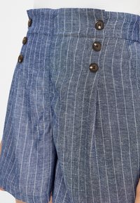 Shorts rayés bleus en tissu texturé avec deux nuances de bleu, trois boutons marron sur la taille, et un détail plissé à l'avant.
