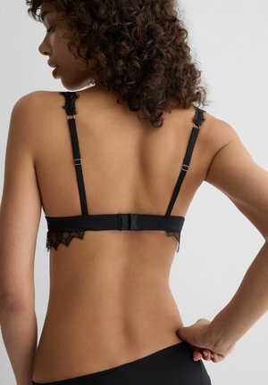 Femme aux cheveux bouclés ajustant la bretelle de son soutien-gorge noir orné de dentelle et son sous-vêtement assorti, vue de dos sur un fond de mur clair uni.