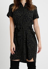 Abito camicia in stampa leopardata verde scuro e nero, maniche corte, chiusura a bottoni sul davanti, vita affusolata con cravatta e spacchi laterali per il movimento.